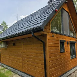 Drewniany dom z czarnym dachem, rynnami i oknami, z anteną satelitarną na dachu, klimatyzacją na ścianie i elementami dekoracyjnymi z drewna.