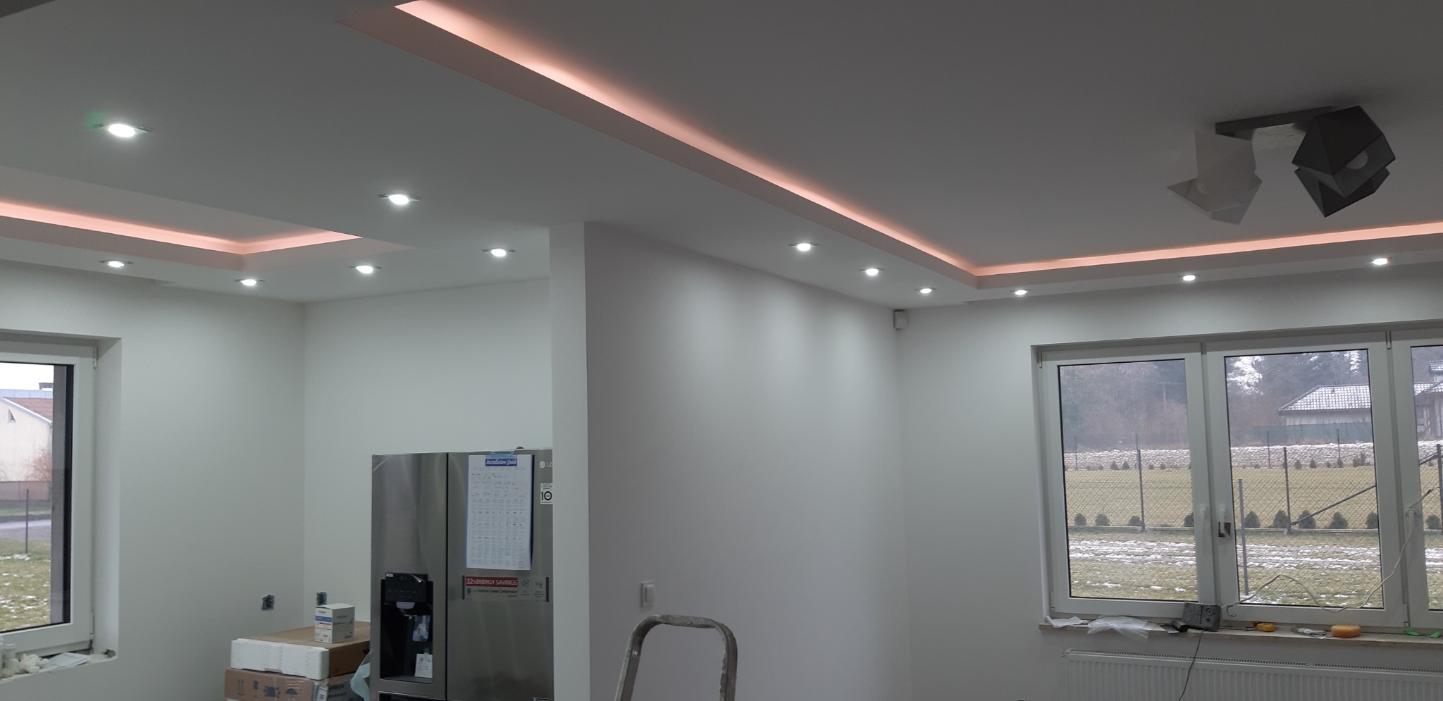 Nowoczesne wnętrze po remoncie: białe ściany, podwieszany sufit z oświetleniem LED, minimalistyczna lampa, widoczne okna i fragment sprzętu AGD.