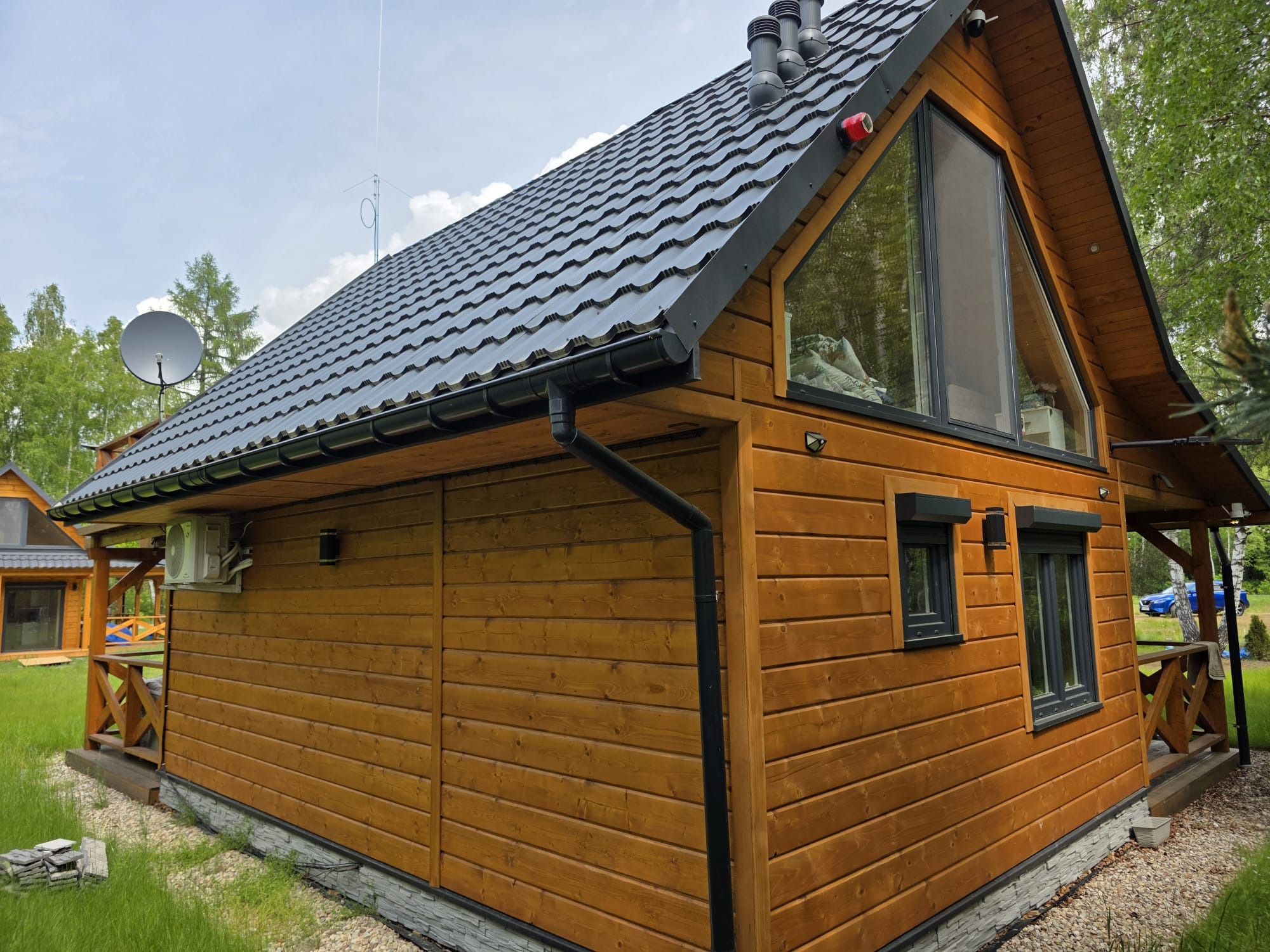Drewniany dom z czarnym dachem, rynnami i oknami, z anteną satelitarną na dachu, klimatyzacją na ścianie i elementami dekoracyjnymi z drewna.