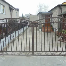 Dwuskrzydłowa brama wjazdowa z metalu, z ozdobnymi elementami, w kolorze ciemnego brązu, z widocznym fragmentem ogrodzenia panelowego po lewej stronie, na tle domów jednorodzinnych.