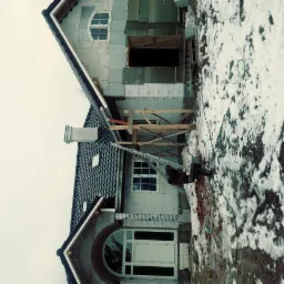 Budowa domu jednorodzinnego w trakcie prac dekarskich, widoczna część dachu pokryta ciemną dachówką, komin, okno dachowe i rusztowanie, na pierwszym planie ośnieżony teren.