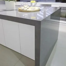 Szary blat kamienny w nowoczesnej kuchni, na blacie dzbanek z wodą i cytryną oraz szklanki na drewnianej tacy, w tle okno z czarnymi ramami.