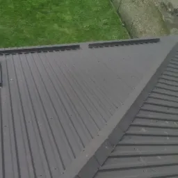 Widok z góry na szary, blaszany dach dwuspadowy z dwoma oknami dachowymi i elementami obróbki blacharskiej na kalenicy, na tle zielonego trawnika i fragmentu budynku.