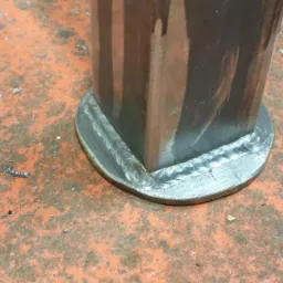 Spoina łącząca kwadratowy profil z okrągłą podstawą, widoczne ślady obróbki metalu i odpryski na pomarańczowym, matowym podłożu. Detal wykonanej spoiny.