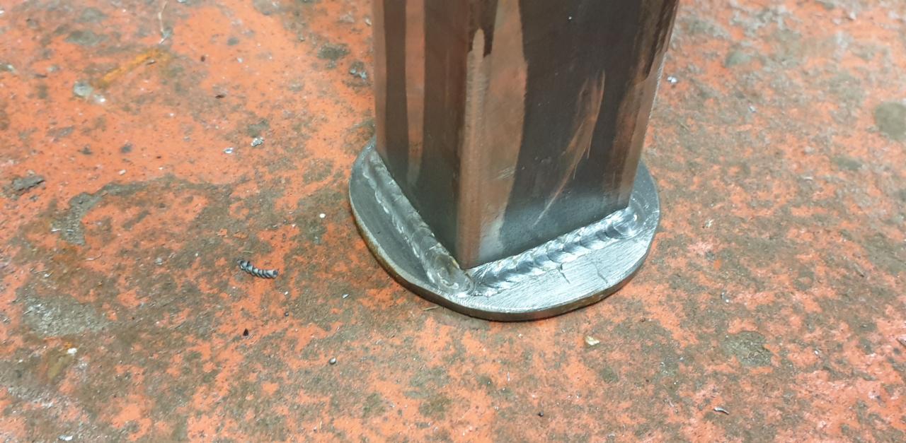 Spoina łącząca kwadratowy profil z okrągłą podstawą, widoczne ślady obróbki metalu i odpryski na pomarańczowym, matowym podłożu. Detal wykonanej spoiny.