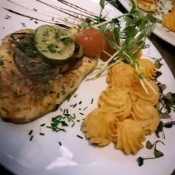 Elegancko podane danie: grillowany filet z kurczaka z karmelizowaną cebulą, ogórkiem, pomidorkami cherry, puree ziemniaczanym i świeżymi ziołami na białym talerzu z dekoracyjnym sosem.