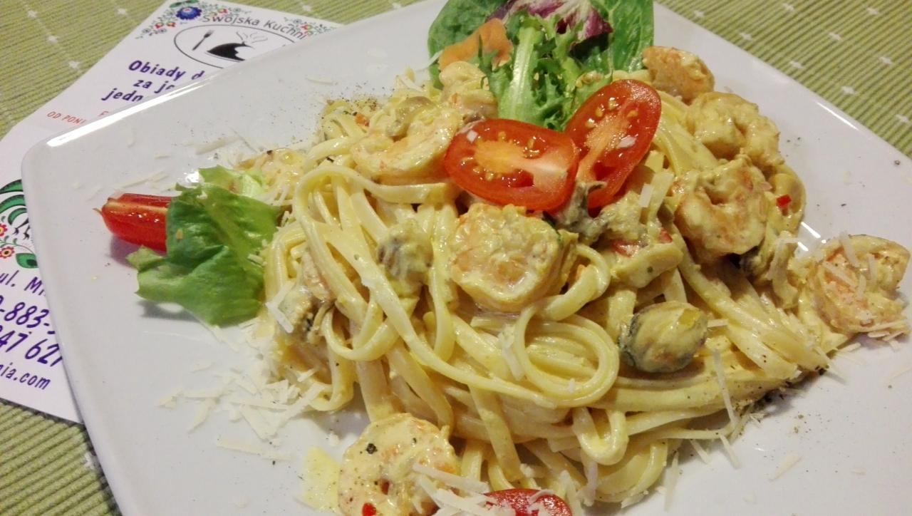 Linguine z krewetkami i małżami w kremowym sosie, udekorowane pomidorkami cherry i sałatą, podane na białym talerzu z ulotką restauracji w tle.