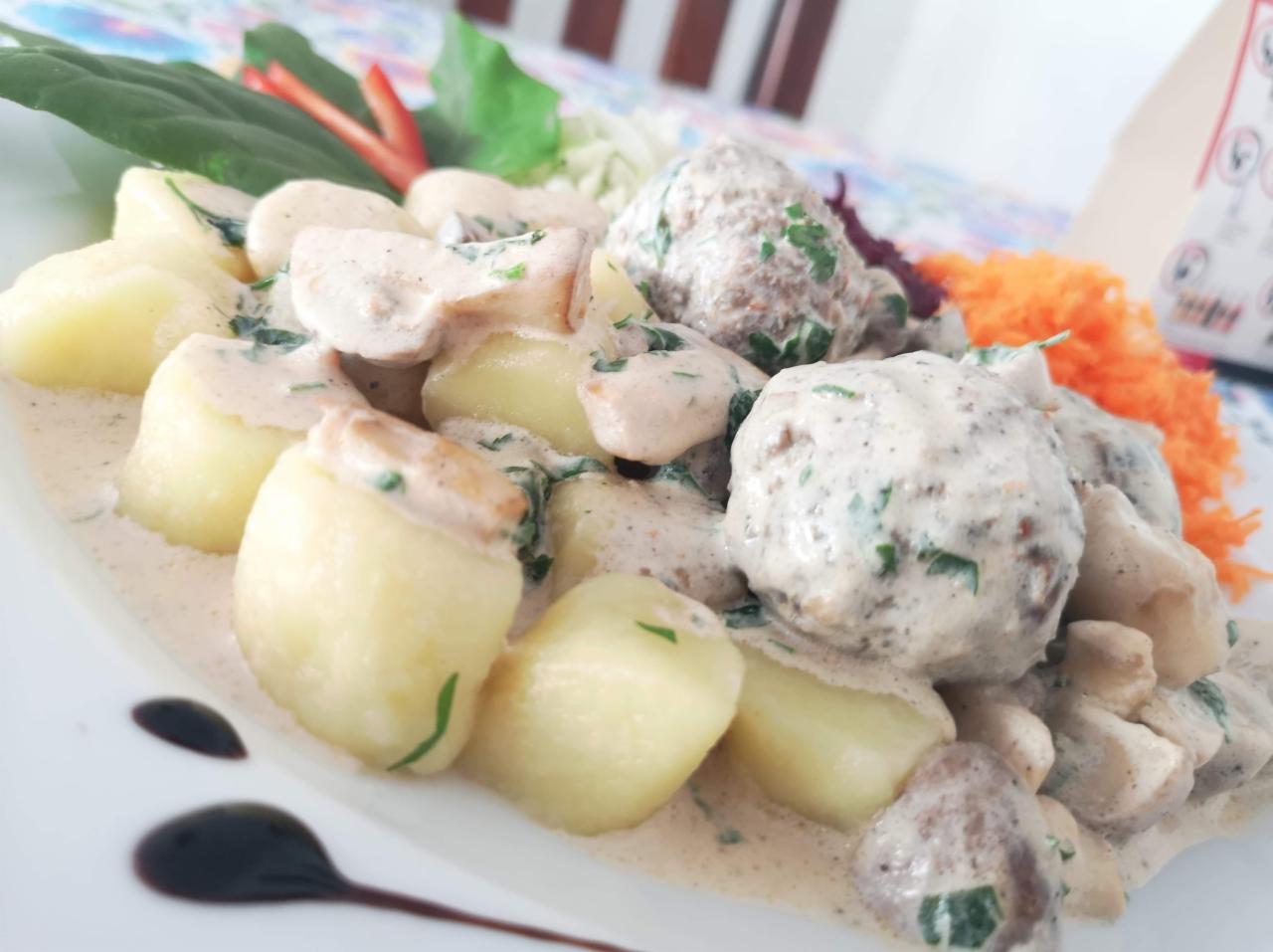 Kluski ziemniaczane gnocchi w kremowym sosie z grzybami i klopsikami, udekorowane świeżymi ziołami i startą marchewką na białym talerzu.