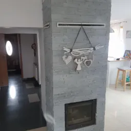 Obudowa kominka z szarej, dekoracyjnej cegły, ozdobiona drewnianą dekoracją z sercami i aniołem, widoczna w jasnym wnętrzu z ciemną podłogą w tle.