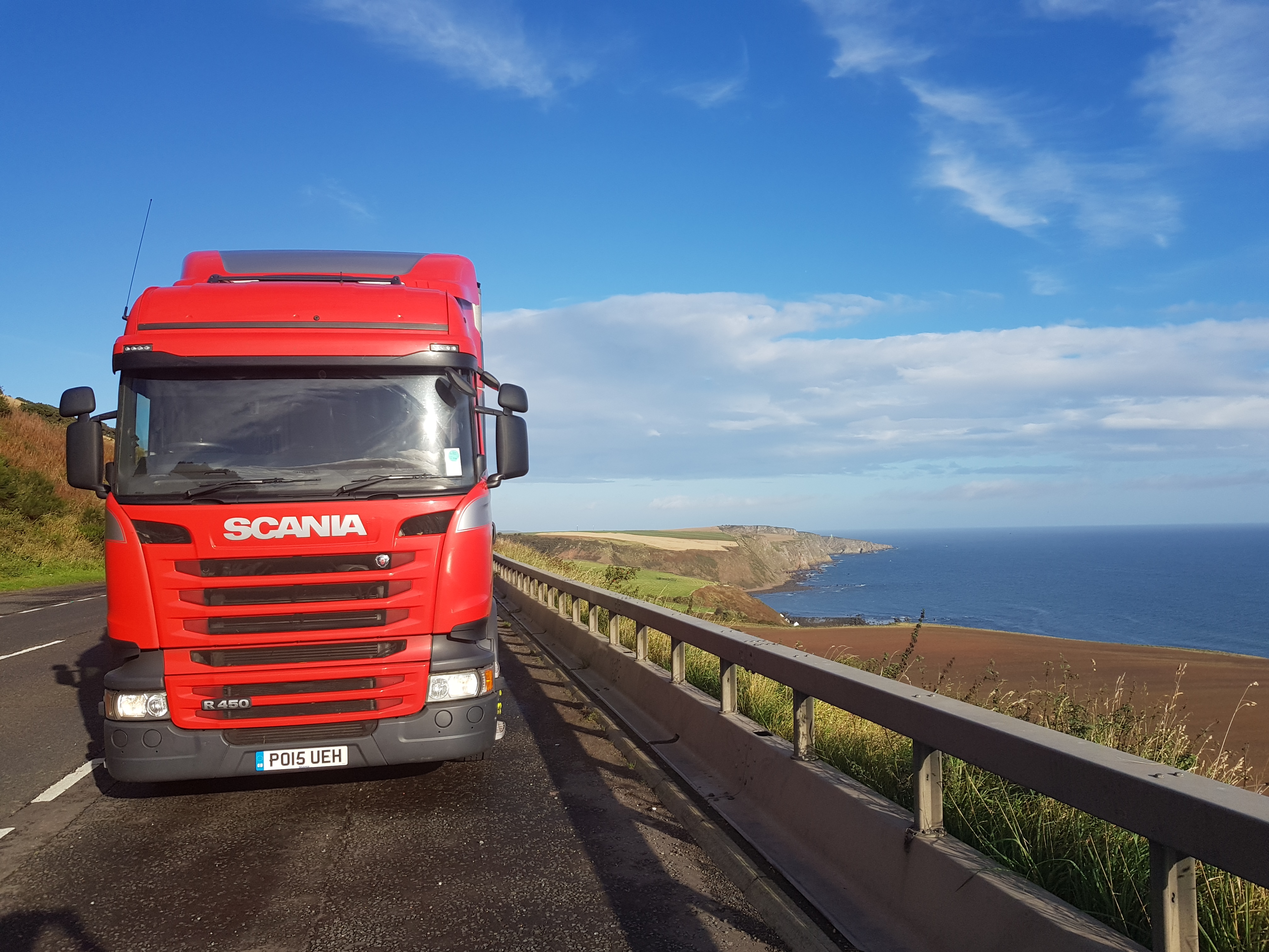 Czerwona ciężarówka Scania R450 na drodze z widokiem na klify i morze w słoneczny dzień.