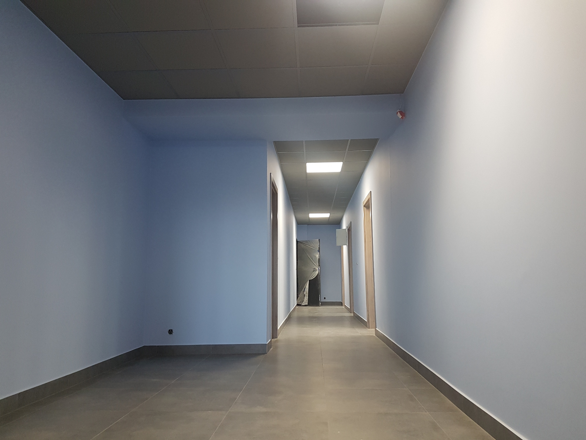 Długi korytarz biurowy z szarymi płytkami na podłodze i świeżo pomalowanymi na jasnoniebiesko ścianami, widoczne drzwi do pomieszczeń wzdłuż korytarza.