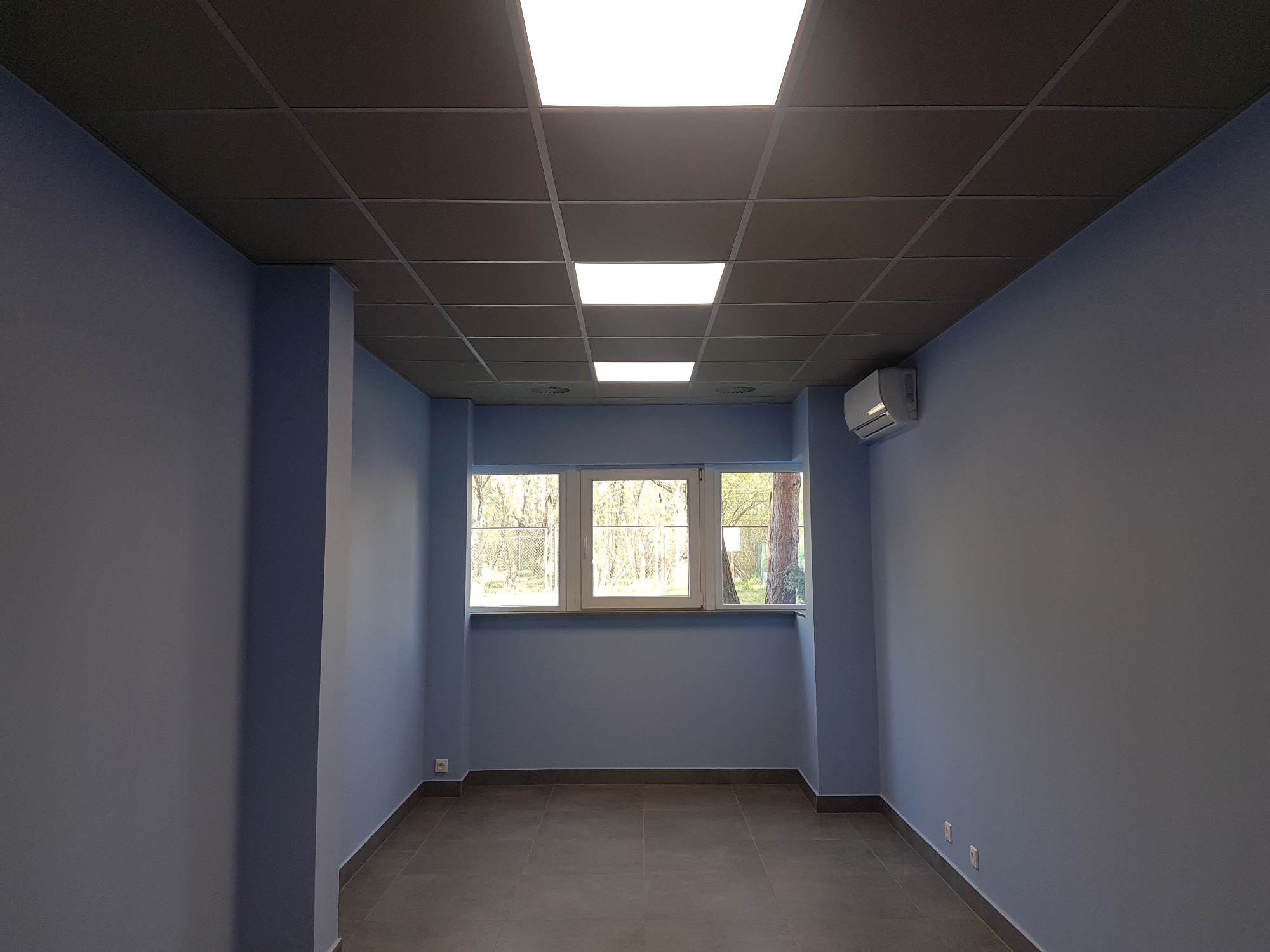 Puste, pomalowane na niebiesko pomieszczenie z trzema oknami, szarą podłogą z płytek i podwieszanym sufitem kasetonowym z wbudowanym oświetleniem LED, klimatyzator na ścianie.