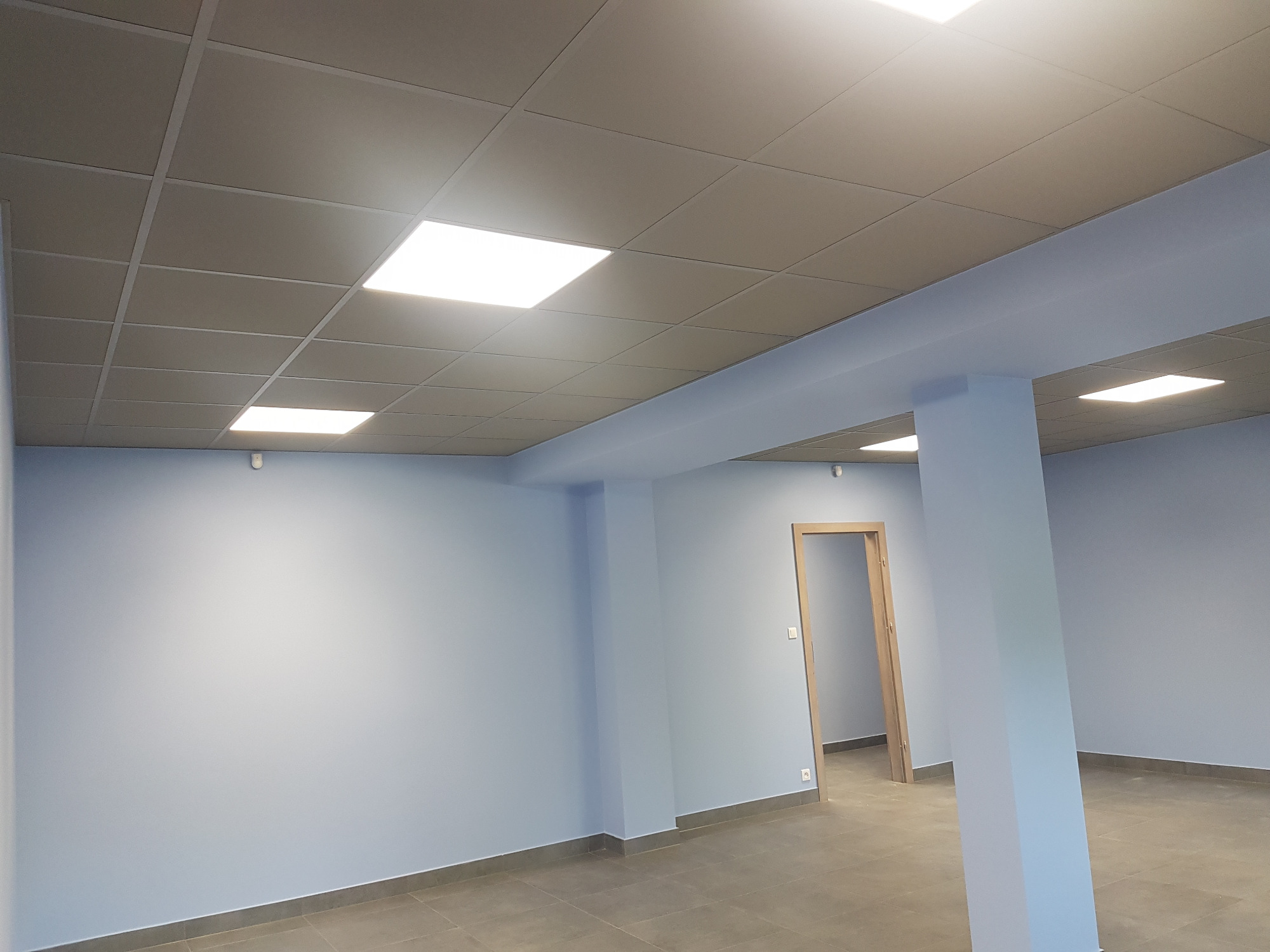 Wnętrze biura po odświeżeniu: świeżo pomalowane ściany w odcieniach błękitu, sufit podwieszany z kasetonami i wbudowanym oświetleniem LED, widoczna kolumna nośna i fragment podłogi z płytek.