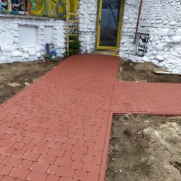 Czerwona kostka brukowa ułożona jako ścieżka prowadząca do dziwacznego domu z żółtymi drzwiami i białymi, okrągłymi dekoracjami na ścianach. Widoczne nierówności terenu po bokach ścieżki.