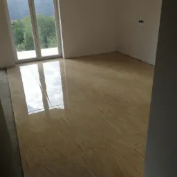 Beżowe, błyszczące płytki ceramiczne ułożone na podłodze w pustym pokoju z dużym oknem, widoczny fragment nieukończonej podłogi przy ścianie.