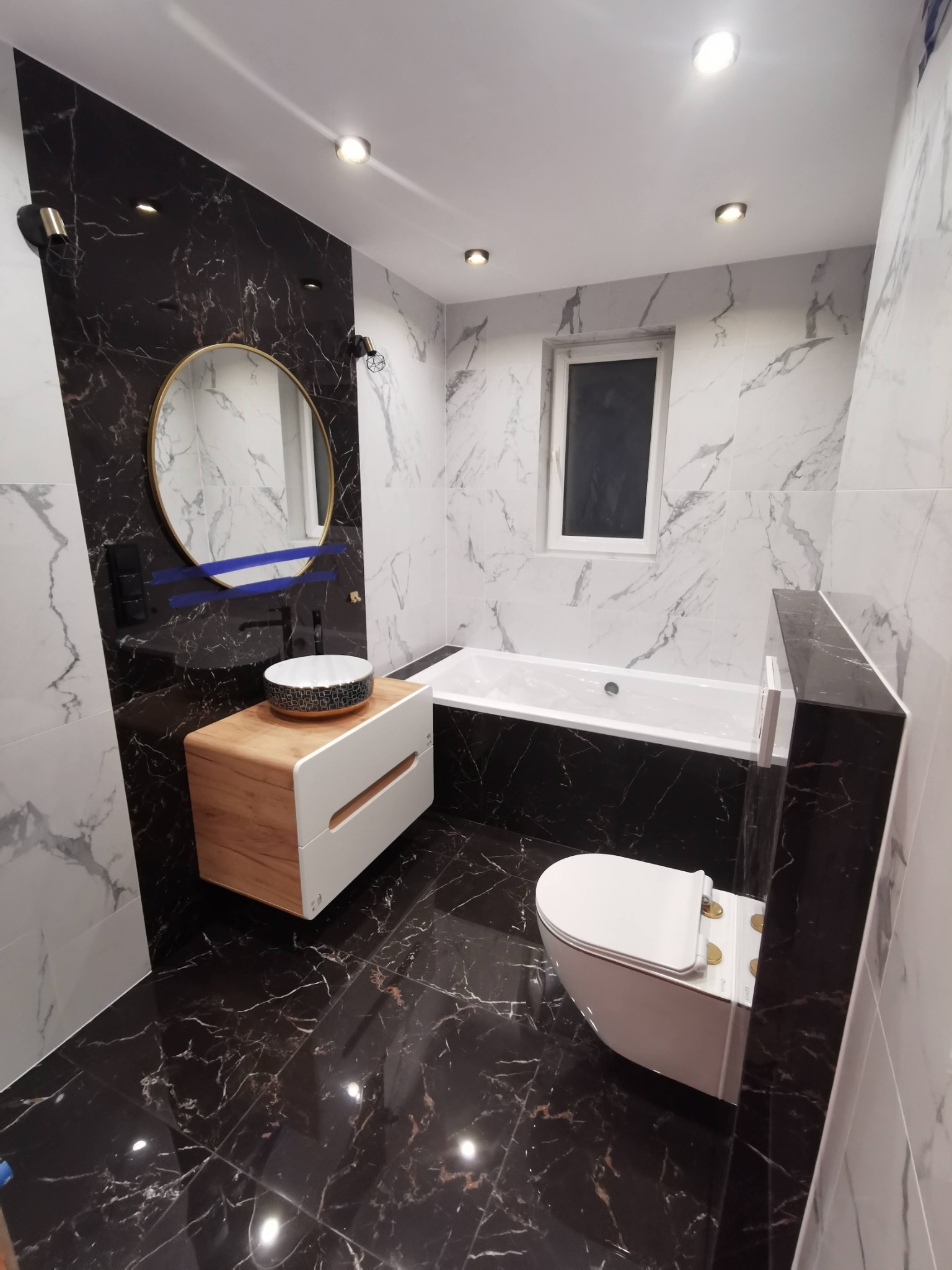 Nowoczesna łazienka z czarno-białymi marmurowymi płytkami, okrągłym lustrem w złotej ramie, umywalką nablatową z dekoracyjnym wzorem i podwieszaną toaletą.