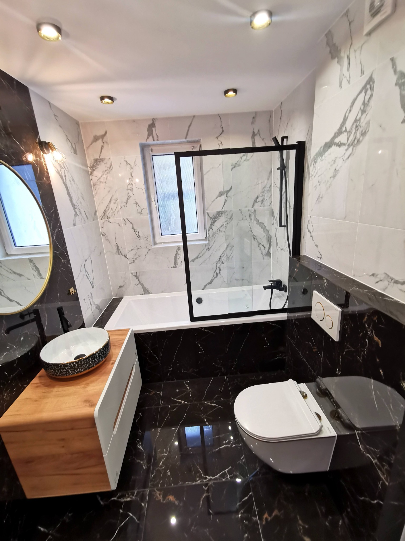 Nowoczesna łazienka z czarno-białymi marmurkowymi płytkami, drewnianą szafką pod umywalkę i czarną armaturą, oświetlona punktowymi lampami sufitowymi.