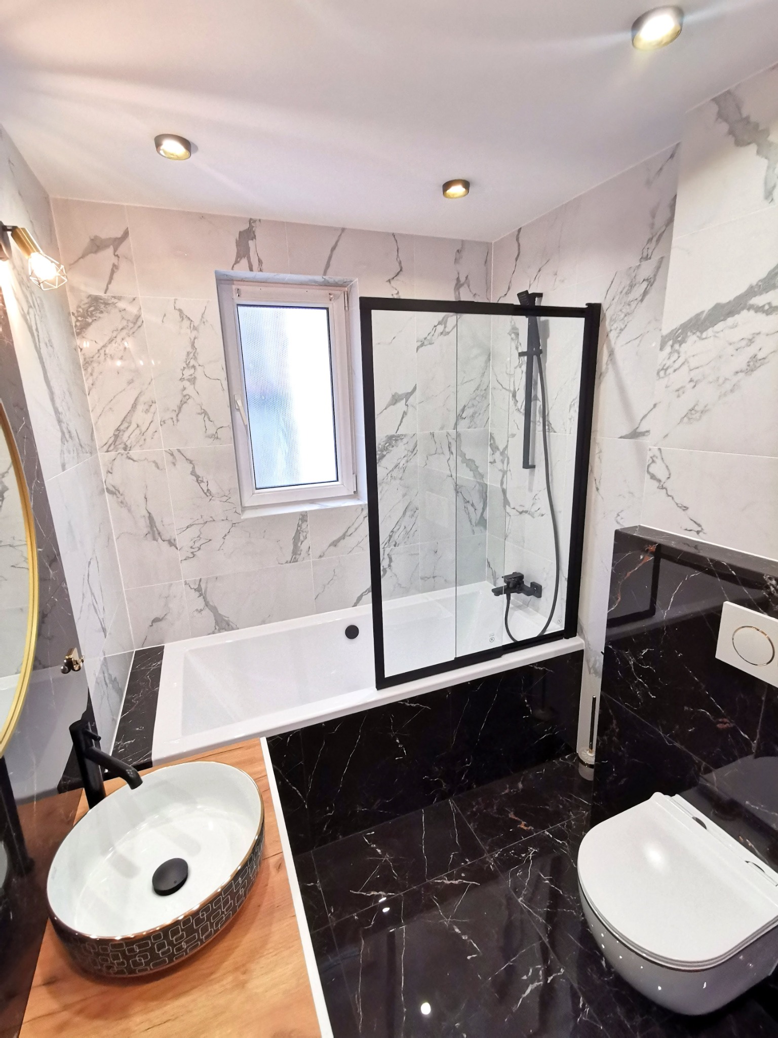 Nowoczesna łazienka z marmurowymi płytkami, czarną armaturą i kabiną prysznicową z czarnymi ramami, owalną umywalką na drewnianym blacie i wiszącą toaletą.