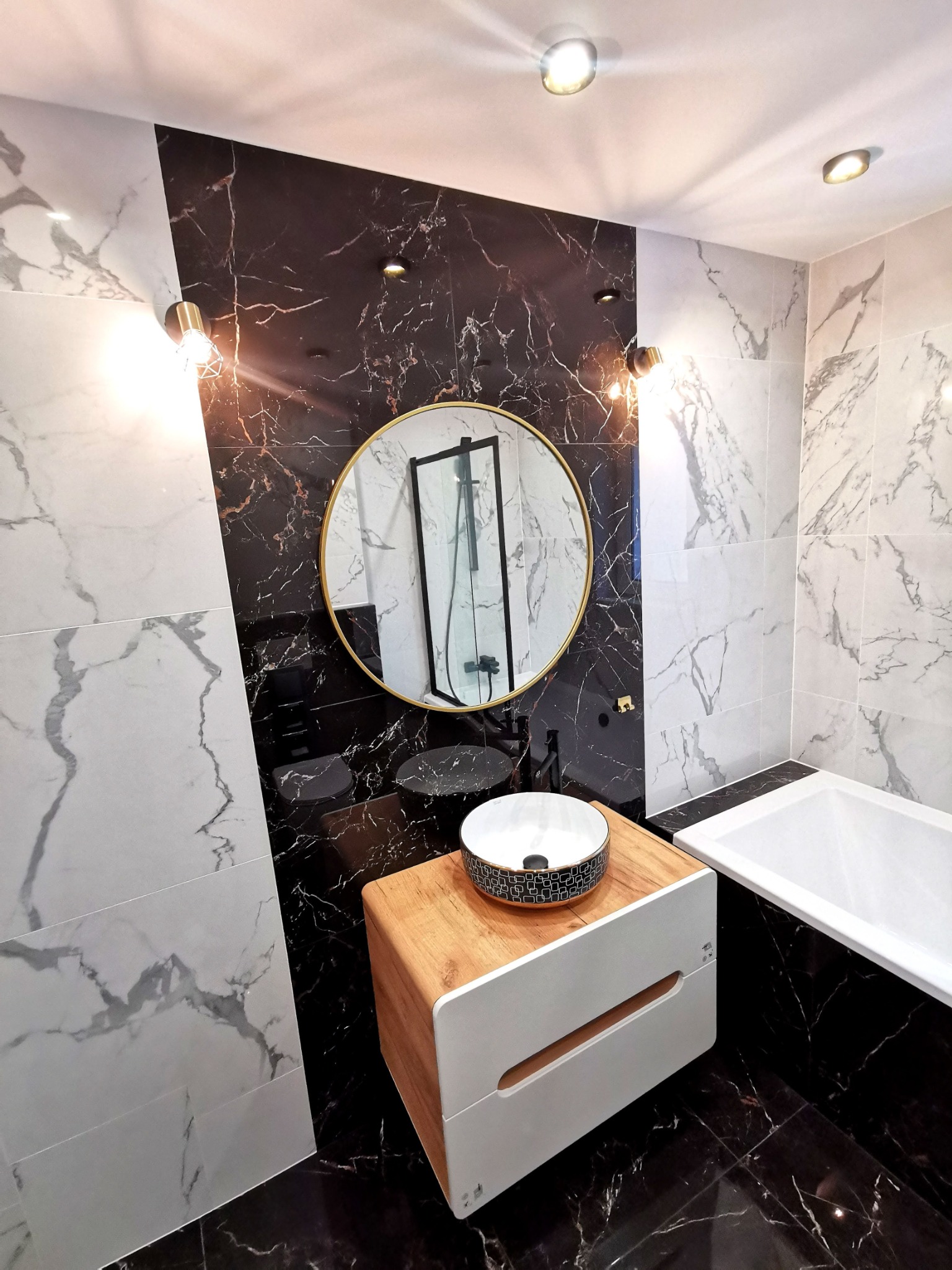 Nowoczesna łazienka z czarno-białymi płytkami imitującymi marmur, okrągłym lustrem w złotej ramie i umywalką nablatową z drewnianym blatem.