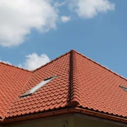 Czerwony dach z dachówki ceramicznej z dwoma oknami dachowymi na tle błękitnego nieba z chmurami.
