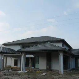 Nowoczesny dom jednorodzinny z szarą dachówką, widoczne rynny, elewacja w jasnym kolorze, teren w trakcie prac wykończeniowych.