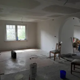 Pokój w trakcie prac wykończeniowych: ściany po nałożeniu gładzi, widoczne niedoskonałości na suficie, przygotowane wiadra z zaprawą, drabina i radio na podłodze.