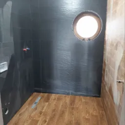 Wnętrze łazienki w trakcie wykańczania, z czarnymi płytkami o fakturze łupka na ścianach, drewnopodobną podłogą, okrągłym otworem okiennym w ścianie i częściowo wykończoną ścianą z płytek...