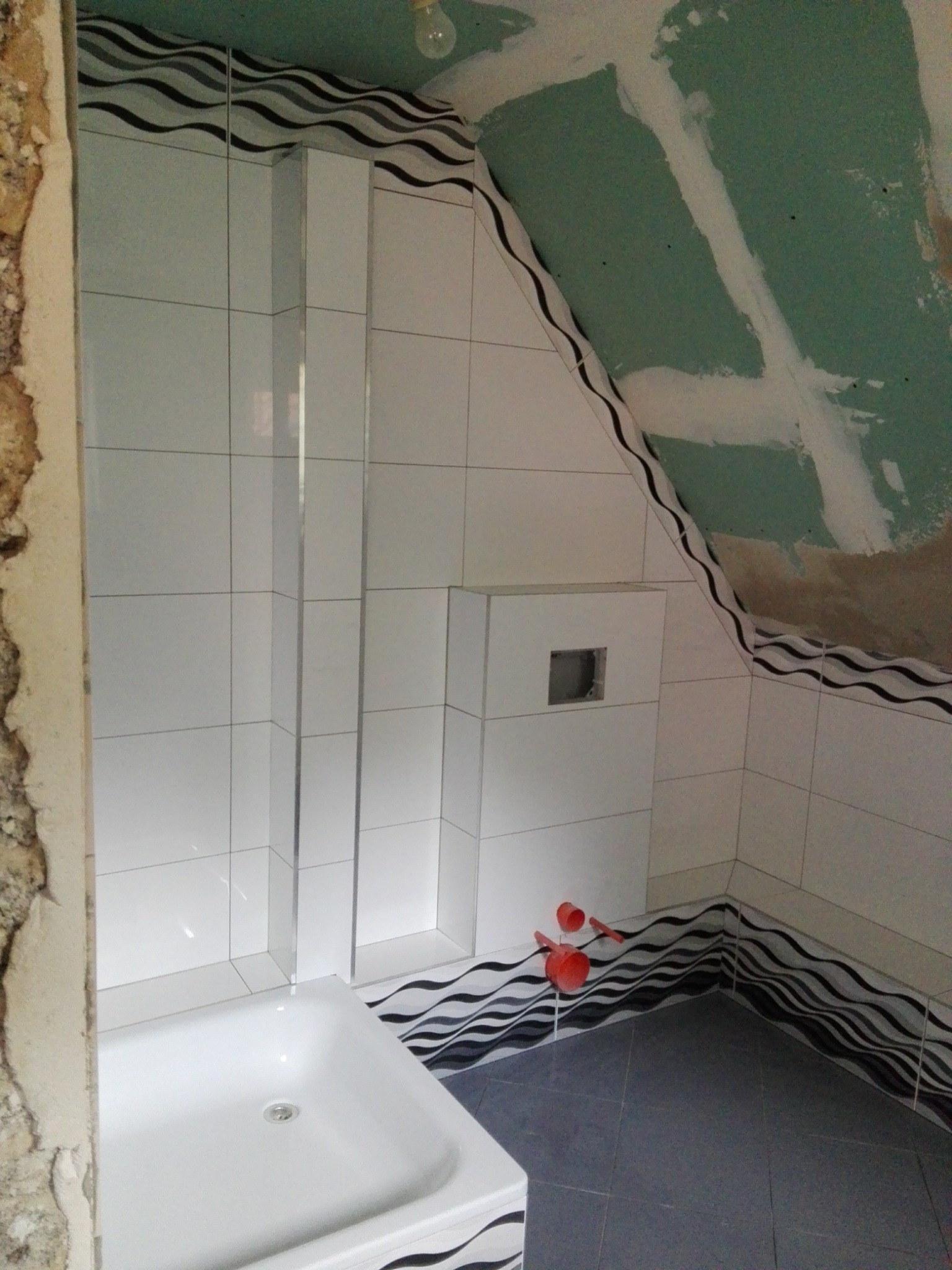 Wnętrze łazienki w trakcie remontu: białe płytki z dekoracyjnym motywem fal, częściowo wykończona ściana z płyty karton gips na poddaszu, widoczna wanna i narzędzia.