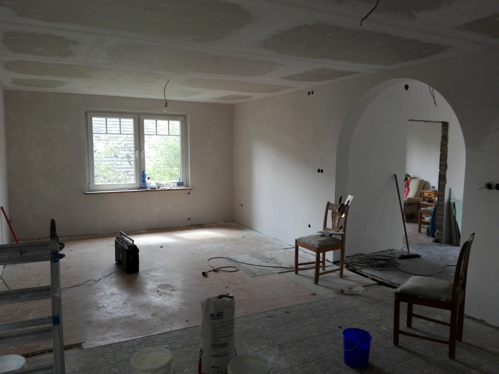 Pokój w trakcie prac wykończeniowych: ściany po nałożeniu gładzi, widoczne niedoskonałości na suficie, przygotowane wiadra z zaprawą, drabina i radio na podłodze.