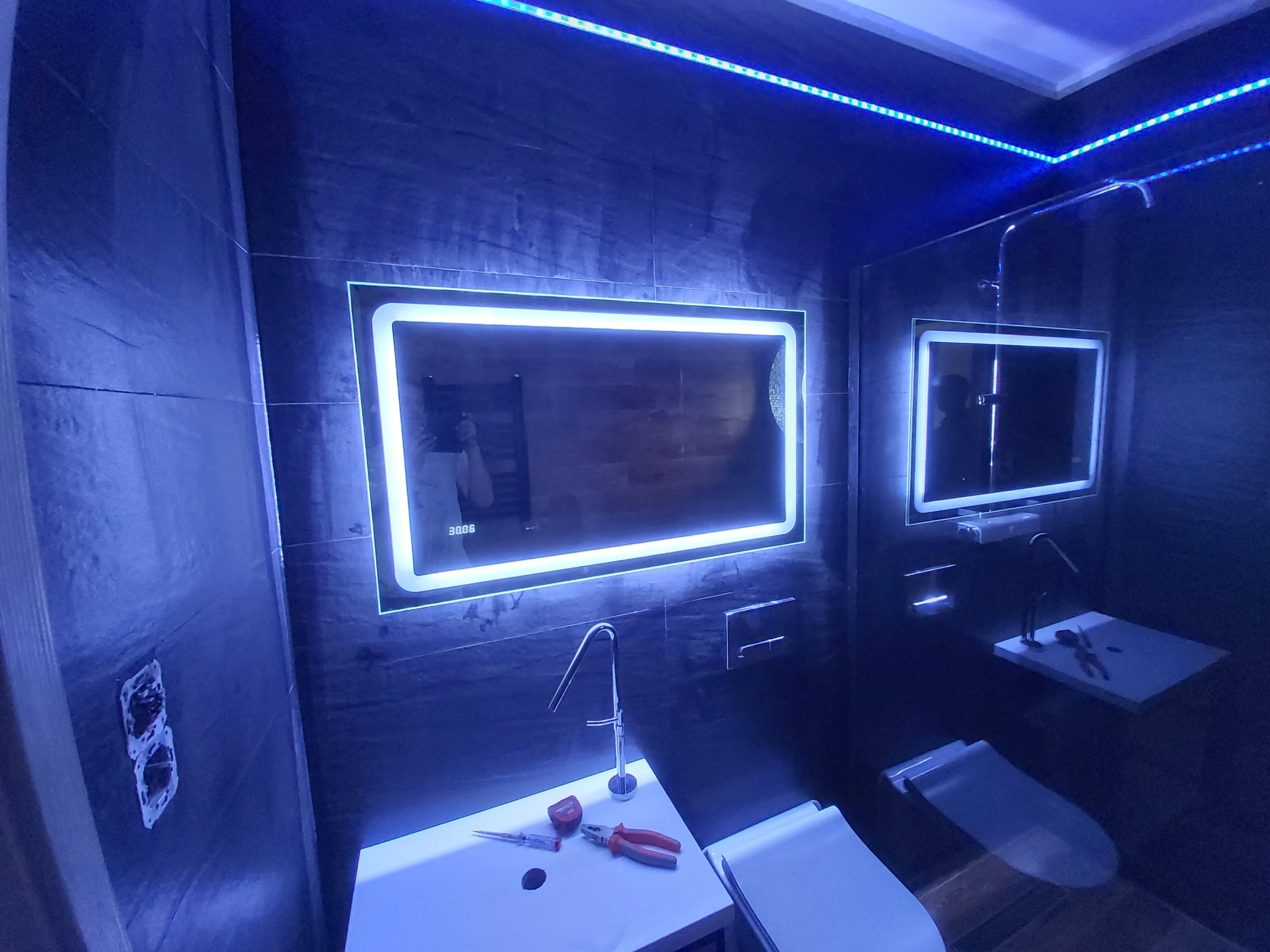 Nowoczesna łazienka z ciemnymi płytkami, podświetlanymi lustrami i ledowym oświetleniem sufitowym; na umywalce leżą narzędzia: wkrętak i kombinerki.