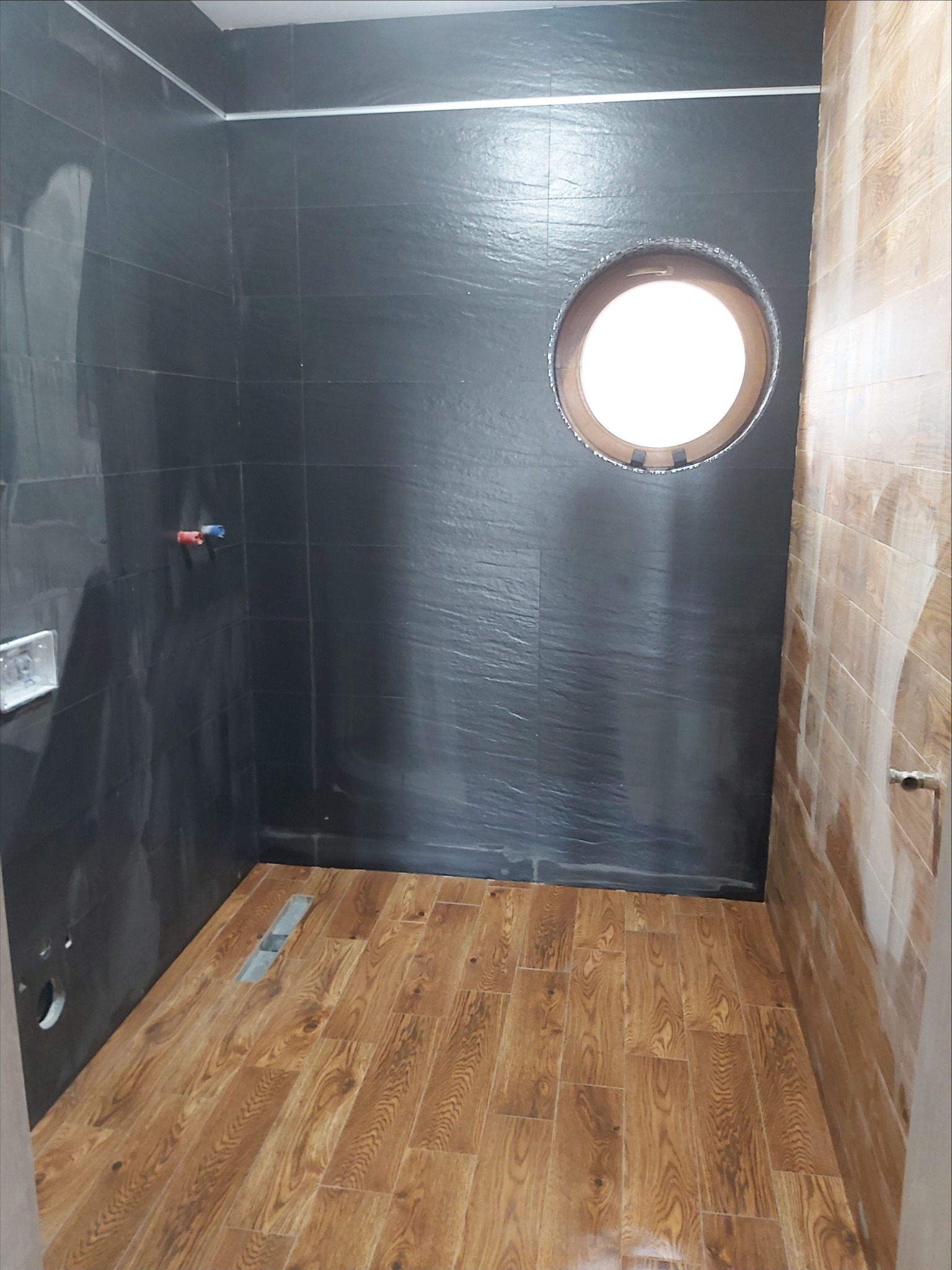 Wnętrze łazienki w trakcie wykańczania, z czarnymi płytkami o fakturze łupka na ścianach, drewnopodobną podłogą, okrągłym otworem okiennym w ścianie i częściowo wykończoną ścianą z płytek...