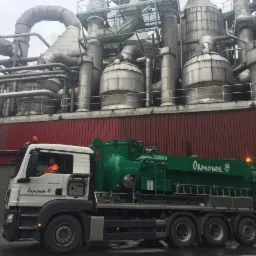 Ciężarówka ssąca z operatorem w pomarańczowej kamizelce odblaskowej na tle instalacji przemysłowej z rurami i zbiornikami.