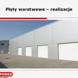 Hala przemysłowa z szarych płyt warstwowych z białymi bramami segmentowymi, widok z zewnątrz, na pierwszym planie kostka brukowa i zwinięty wąż.