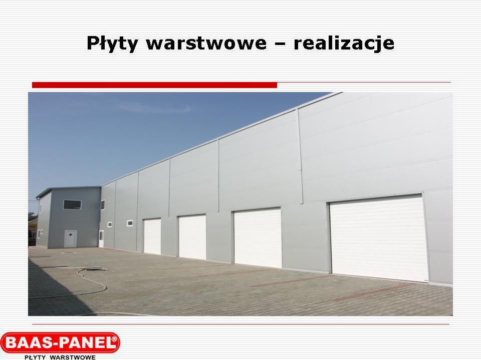 Hala przemysłowa z szarych płyt warstwowych z białymi bramami segmentowymi, widok z zewnątrz, na pierwszym planie kostka brukowa i zwinięty wąż.