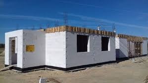 Wznoszenie ścian parterowego budynku z białych bloczków, widoczne szalunki pod wieniec i wystające pręty zbrojeniowe, błękitne niebo w tle.