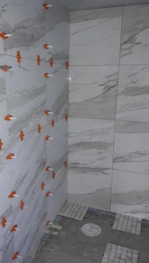 Ściana łazienki w trakcie układania płytek imitujących marmur, z widocznymi pomarańczowymi krzyżykami dystansowymi i systemem poziomowania, na podłodze fragment ułożonej mozaiki.