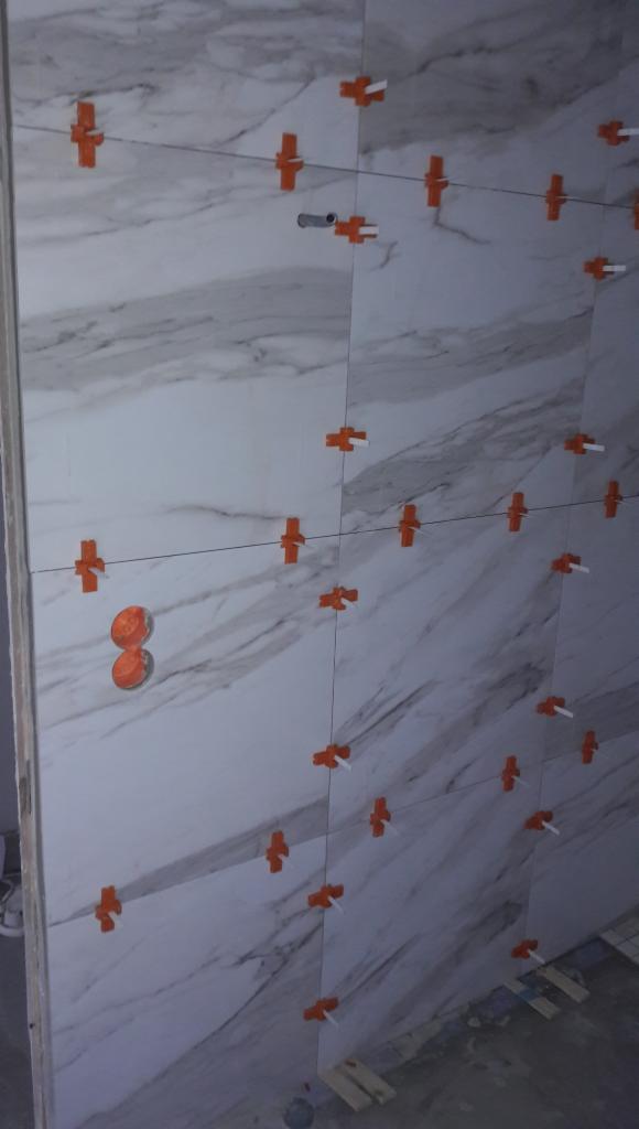 Układanie marmurowych płytek na ścianie w Warszawie, z użyciem pomarańczowych krzyżyków dystansowych i systemu poziomowania, widoczne gniazdka elektryczne.