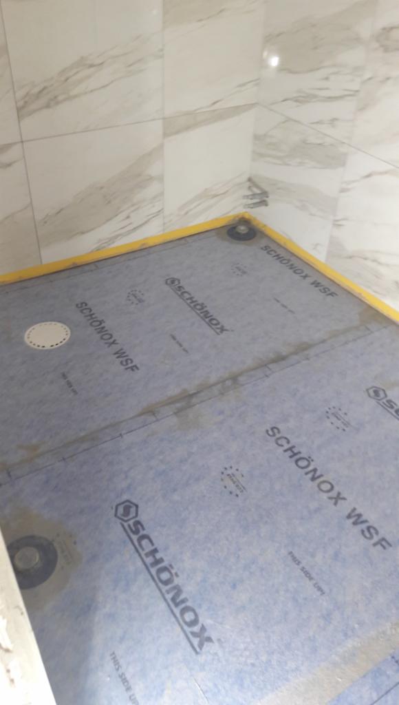 Przygotowanie podłoża pod prysznic z użyciem membrany hydroizolacyjnej SchönoX WSF, widoczne łączenia zabezpieczone żółtą taśmą, marmurowe płytki na ścianach.