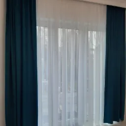 Okno z białymi firanami i zasłonami w kolorze butelkowej zieleni, widoczne fałdy materiału.