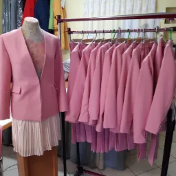 Różowy damski garnitur na manekinie krawieckim, z rzędem identycznych garniturów na wieszakach w tle, w pracowni.