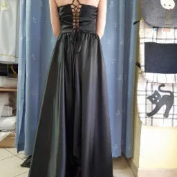 Tył czarnej, satynowej sukni wieczorowej z wiązaniem na plecach, widoczny na tle błękitnej zasłony z delikatnym wzorem i półki z tkaninami w pracowni.