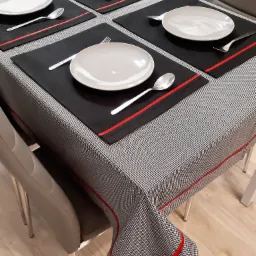 Nakryty stół z czarno-białym obrusem w pepitkę, czarnymi podkładkami z czerwoną lamówką, talerzami i sztućcami.