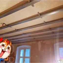 Montaż metalowej konstrukcji podwieszanego sufitu w pomieszczeniu z oknami, widoczne profile i płyty. Na pierwszym planie naklejka.