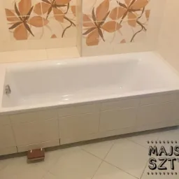 Biała wanna obudowana płytkami w jasnej łazience z beżowymi kwiatowymi płytkami dekoracyjnymi na ścianie. Mały drewniany element pod wanną.