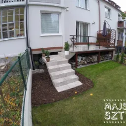 Widok z zewnątrz na dom z tarasem, schodami wyłożonymi płytkami, trawnikiem i ogrodzeniem z siatki; pod tarasem składowane drewno.