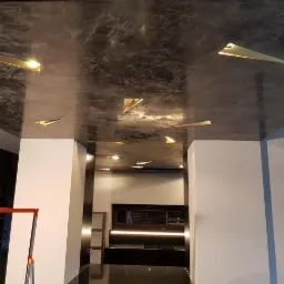 Szary sufit z efektem betonu architektonicznego i złotymi, trójkątnymi lampami LED w nowoczesnym wnętrzu, widoczna drabina malarska i fragment zabudowy kuchennej.