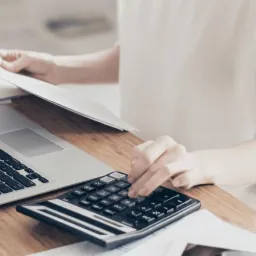 Kobieta w jasnej bluzce używa kalkulatora obok otwartego laptopa i sterty dokumentów na drewnianym biurku.