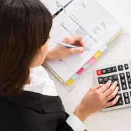 Kobieta w czarnej marynarce, odwrócona tyłem, wypełnia dokumenty w segregatorze, używając srebrnego długopisu, obok kalkulator z wyświetlonym wynikiem na białym biurku.