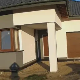 Nowoczesny dom jednorodzinny z kremową elewacją, szarymi akcentami i brązową bramą garażową, widok z zewnątrz na tle błękitnego nieba i zieleni drzew.