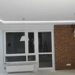 Wnętrze pokoju z białymi oknami i grzejnikiem, ceglana ściana, sufit z oświetleniem LED i wisząca lampa.